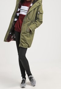 Oliwkowy parka z futrzanym kapturem i elastycznym pasem, noszona na bordowym swetrze i czarnych dopasowanych dżinsach, w zestawieniu z czarno-białymi trampkami.