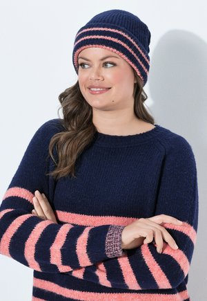 Frau trägt eine marineblaue und pink gestreifte Strickmütze und passenden Pullover, hat die Arme verschränkt, lächelt und schaut zur Seite.