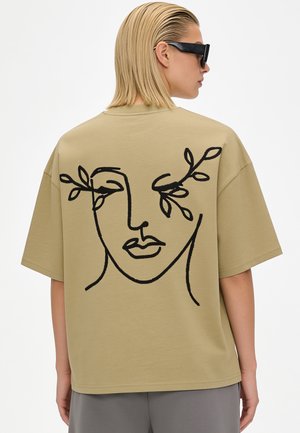 Person trägt ein hellbraunes oversized T-Shirt mit schwarzem abstraktem Gesichts- und Blattdesign auf dem Rücken und schwarze Sonnenbrille, steht seitlich.