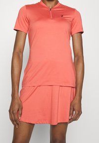 Polo shirt atletico corallo con una breve cerniera, abbinato a una gonna svasata coordinata. Realizzato in tessuto leggero e liscio. Dettaglio del logo semplice.