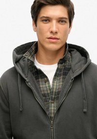 Sudadera gris con cremallera y capucha con cordón, superpuesta sobre una camisa de cuadros verde. La sudadera presenta una textura suave y un corte casual.