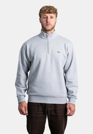 Mann mit lockigem Haar trägt einen hellgrauen Reißverschluss-Pullover und dunkle Hose, steht vor einem einfarbigen hellen Hintergrund und blickt mit neutralem Ausdruck nach vorne.