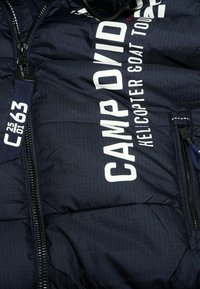 Donkerblauwe gewatteerde jas met witte tekst "CAMP DAVID", ondertitel "Helicopter & Coat Tour" en ritssluiting met label "C63 25 01" aan de voorkant.