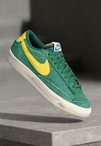 Zelene nubuk tene s prašnato rumenim Nike znakom, z belo gumijasto podplatom in teksturiranimi detajli. Ima klasičen oblikovanje s vezalkami.