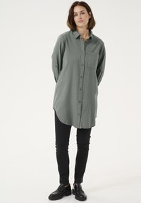 Pikkade varrukatega, nööpidega rohekas-halli tooni särk, lõdva lõike ja ümarate äärtega, ühe rinnataskuga, kokku sobitatud mustade kitsaste teksade ja mustade kingadega.