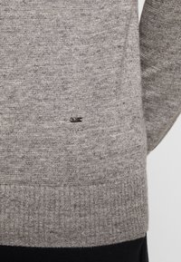 Grå stickad tröja med ribbad nederkant och en liten svart logotypdetalj nära botten. Mjuk textur med ett enhetligt mönster.