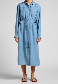 Nainen, joka on pukeutunut vaaleansiniseen denim-mekkoon, jossa on pitkä hihat, napit edessä ja vyötärövyö, sekä beigeihin avonaisiin sandaaleihin.