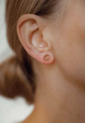 Goldenes Friedenszeichen-Stift-Ohrstecker mit kreisförmigem Design und durchbrochenem Symbol. Glatte Oberfläche, minimalistischer Stil, wird im Ohrläppchen getragen.