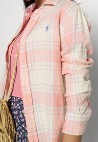 Camisa de cuadro ligera en rosa y crema. Presenta un cierre de botones en el frente, mangas enrolladas y un detalle de logo azul en el pecho.