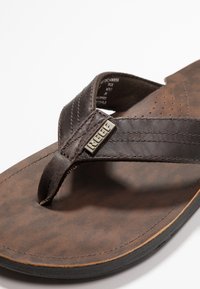 Infradito in pelle marrone con soletta testurizzata, cinturini cuciti e un accento in metallo con logo. Suola piatta con leggera curvatura.