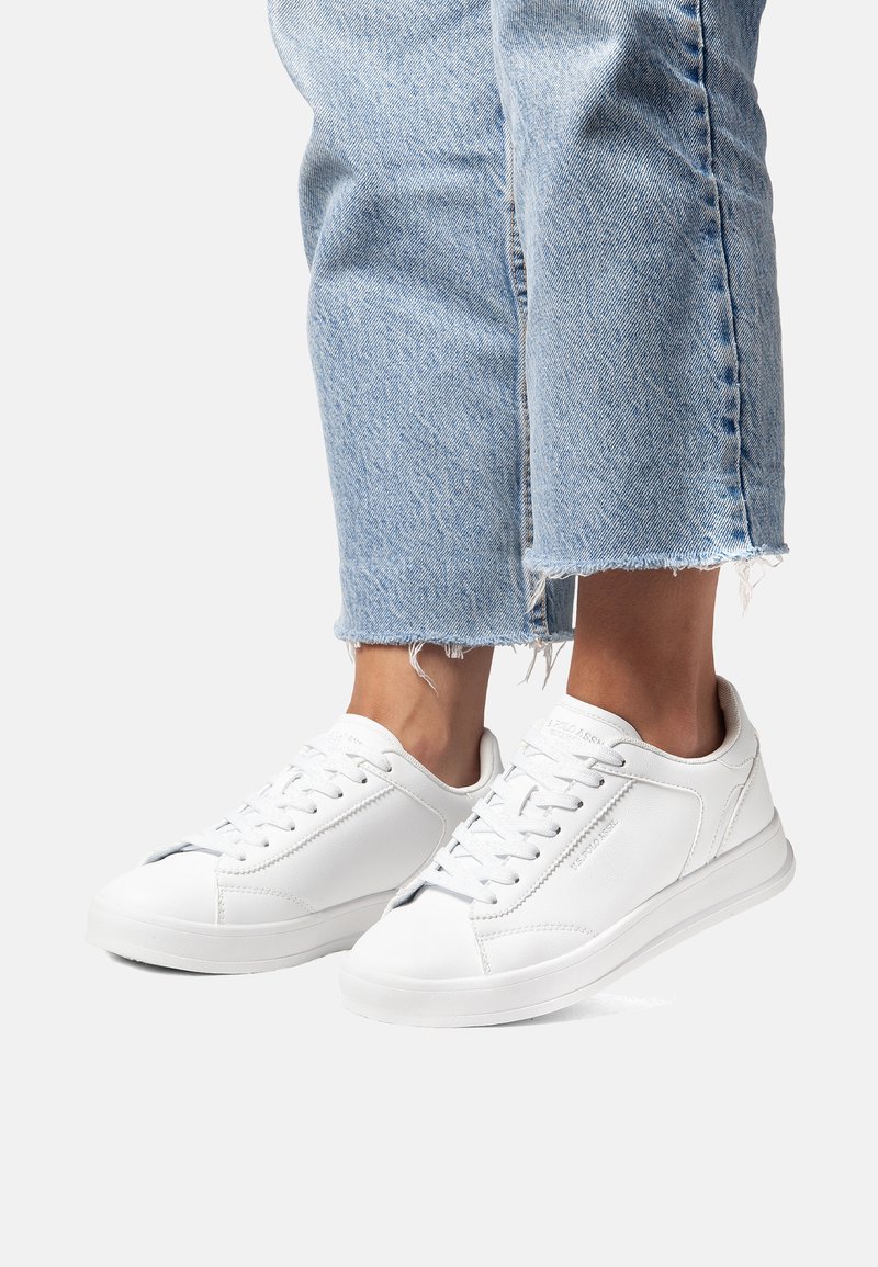 White sneakers made of smooth leather with a lace-up front, rounded toe, and subtle stitching. Paired with light blue frayed hem jeans.