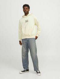 Creme hættetrøje med en kængurulomme og logo-print, parret med lyseblå jeans og sorte sneakers, stående foran en lys baggrund.