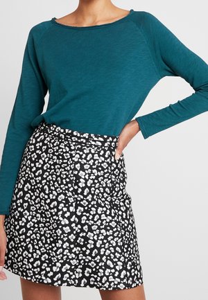 Langærmet t-shirt i blågrøn farve kombineret med et højtaljet nederdel med sort-hvid leopardprint, hånden hviler på hoften.