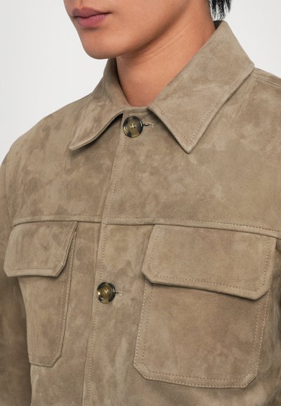Tan suede shirt jacket featuring a pointed collar, two chest pockets, and black buttons with a glossy finish. Soft texture with a matte look.