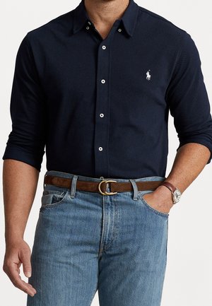 Uomo che indossa una camicia blu navy a maniche lunghe con logo bianco, jeans azzurri, cintura marrone e orologio da polso con cinturino in pelle.