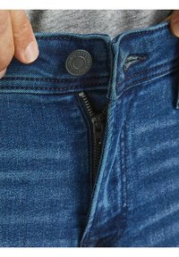 Vaqueros de denim azul oscuro con una cremallera parcialmente subida, que cuentan con un botón de metal, detalles de costura contrastantes y una textura suave cerca de la cinturilla.