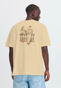 Beige katoenen t-shirt met een geprinte illustratie van een stel dat tegen een reling leunt, met een casual pasvorm en korte mouwen.