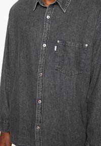 Camicia in denim nero con maniche lunghe, chiusura a bottoni, tasca sul petto e dettagli di cucitura in contrasto. Il tessuto appare morbido con una leggera texture.