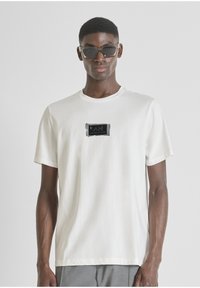 Camiseta de algodón blanca con un parche de logo rectangular negro en el pecho. Manga corta, cuello redondo, textura suave y corte relajado.