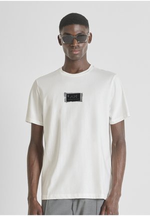 Camiseta de algodón blanca con un parche de logo rectangular negro en el pecho. Manga corta, cuello redondo, textura suave y corte relajado.