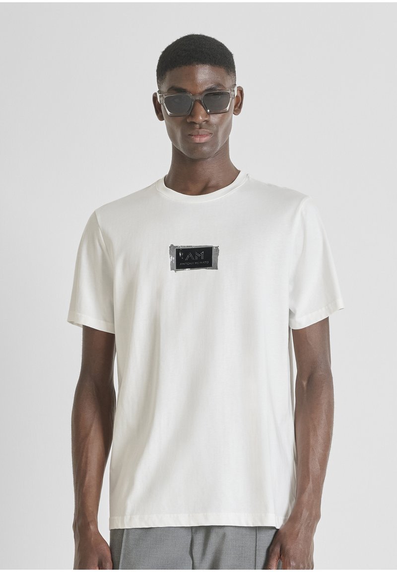 Camiseta de algodón blanca con un parche de logo rectangular negro en el pecho. Manga corta, cuello redondo, textura suave y corte relajado.