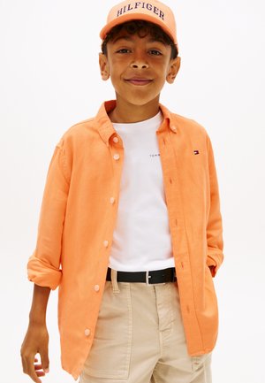 Dreng iført orange Hilfiger kasket og skjorte over hvid t-shirt, beige bukser, smilende med hænder afslappet ned langs siderne mod hvid baggrund.