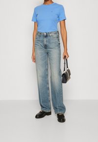 Kvinde iført en lys blå tætsiddende t-shirt, højtaljede bredbenede blege jeans, sorte loafers, og holder en lille sort håndtaske i højre hånd.
