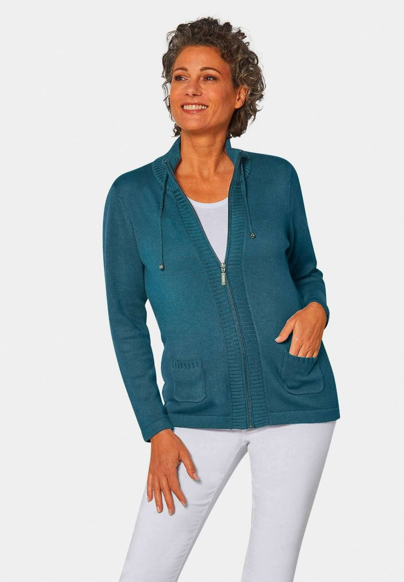 Türkise Reißverschluss-Kardigan mit Kapuze, geripptem Design und zwei frontalen Taschen, kombiniert mit einem weißen Oberteil und hellen Hosen.