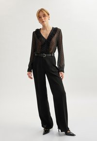 Sort gennemsigtig bluse med polka prikker og bølget krave, samt high-waisted sorte bukser og spidse sorte hæle.