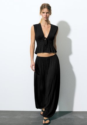 Femme aux cheveux blonds portant un crop top noir sans manches avec un détail noué et un pantalon noir ample, debout contre un mur blanc.