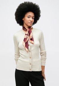 Mulher com penteado afro vestindo um cardigan bege com textura canelada e botões dourados, um lenço de seda em tons de bordô e pêssego, e calças pretas, posando contra um fundo branco.