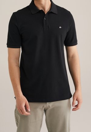 Mann trägt schwarzes kurzärmeliges Poloshirt mit kleinem weißem Sticklogo auf der Brust und beigen Hosen, steht vor einfarbigem Hintergrund.