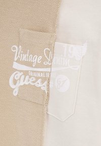 Beige strukturerad tyg med en vit etikett som visar "Vintage Denim" och "Guess" på vänster ficka. Slät vit tyg på höger sida.