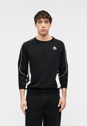 Ung mand med krøllet hår iført en sort Kappa-sweatshirt med pink og hvide stribedetaljer, stående foran en ensfarvet lys baggrund.