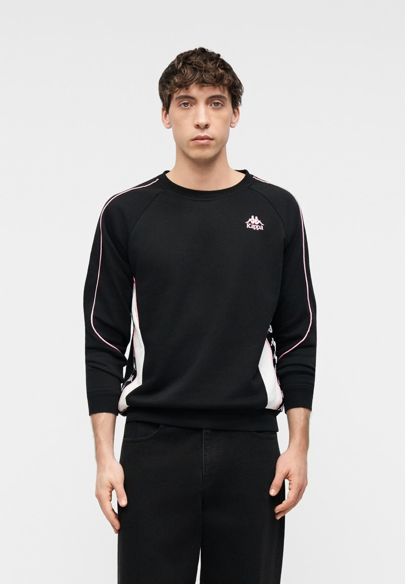 Νέος με σγουρά μαλλιά που φοράει μαύρο φούτερ Kappa με ροζ και λευκές ρίγες, στέκεται μπροστά σε ένα απλό φωτεινό φόντο.