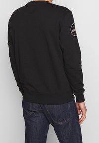 Sudadera negra con cuello redondo, puños y dobladillo acanalados. Presenta un logo de NASA bordado en la manga izquierda. Textura suave.