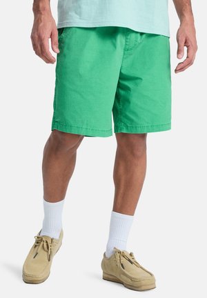 Mann trägt grüne knielange Shorts, weiße Sportsocken und beige lässige Schnürschuhe, steht vor weißem Hintergrund.