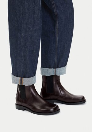 Braune Leder-Stiefeletten mit elastischen Seiteneinsätzen, abgerundetem Zehenbereich und niedrigem Blockabsatz, kombiniert mit dunkelblauen Jeans, die am Saum hochgekrempelt sind.