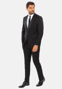 Completo nero con rever in raso, camicia bianca e cravatta nera. Pantaloni slim-fit e scarpe nere lucide. Linee pulite e design su misura.