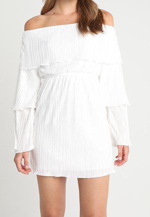 Femme portant une mini-robe blanche plissée, longue manches tombantes, avec des détails à volants sur la poitrine et les manches.