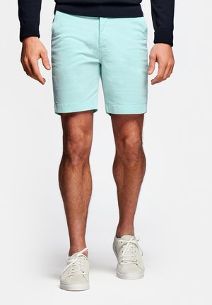 Ben van een persoon die lichtblauwe broekjes, witte sneakers en een zwart shirt met lange mouwen draagt, staand op een witte achtergrond.