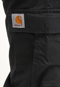 Juodos audinio kargo kelnės su priekinėmis kišenėmis ir baltu etiketė su oranžiniu logotipu, turinčios tinklelio tekstūrą ir tvirtą siuvimą.