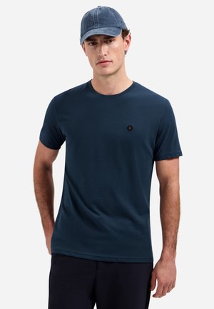 Mann trägt ein marineblaues T-Shirt mit kurzen Ärmeln und eine dunkelblaue Kappe, steht mit den Händen seitlich an einer einfarbigen Wand.