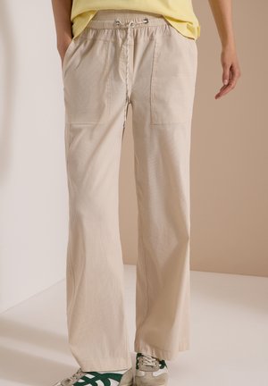 Personne portant un pantalon beige à jambes larges avec poches avant et taille à cordon, associée à des baskets blanches et vertes.