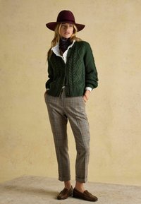 Cardigan verde cu model împletit și nasturi, cămașă albă cu volane, pantaloni în carouri și pantofi maro loafer; asortate cu o pălărie bordeaux cu boruri largi.