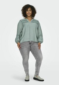 Blusa verde chiaro con scollatura a V, tessuto testurizzato e maniche a palloncino. Abbinata a jeans grigi e mocassini grigio chiaro.