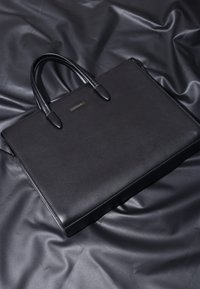 Sort lædermappe med tekstureret overflade, dobbelte håndtag og lynlåslukning. Har en subtil logo-plade på forsiden.