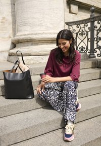 Borsa a mano nera in pelle accanto a una persona con un top bordeaux e pantaloni a palazzo con stampa leopardata, seduta sui gradini di pietra. Tazza di caffè in mano.