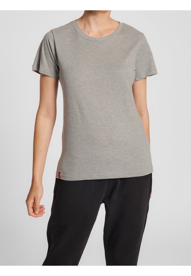 Femme portant un t-shirt gris clair uni à manches courtes et un pantalon noir, debout bras détendus le long du corps sur un fond neutre.