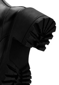 Bota de cuero negro con una suela de goma gruesa que presenta un patrón de tacos resistente para tracción. Textura suave y diseño de punta redonda.